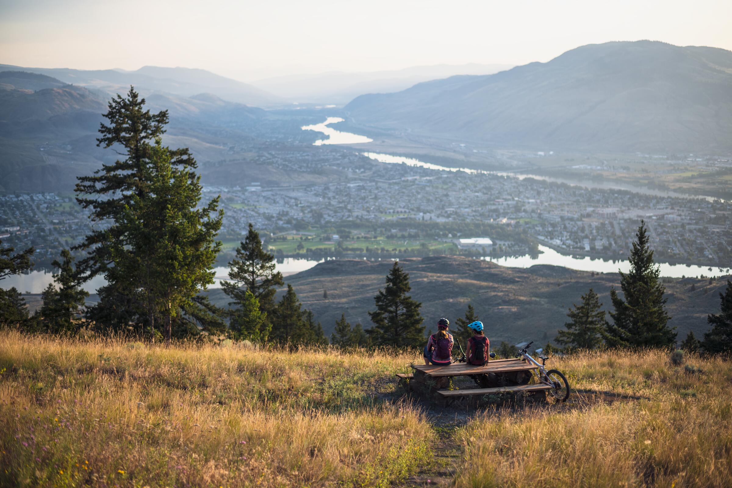 Two riders at a picnic table on a grassy hillside above Kamloops and the Thompson River, a bike leaning beside them