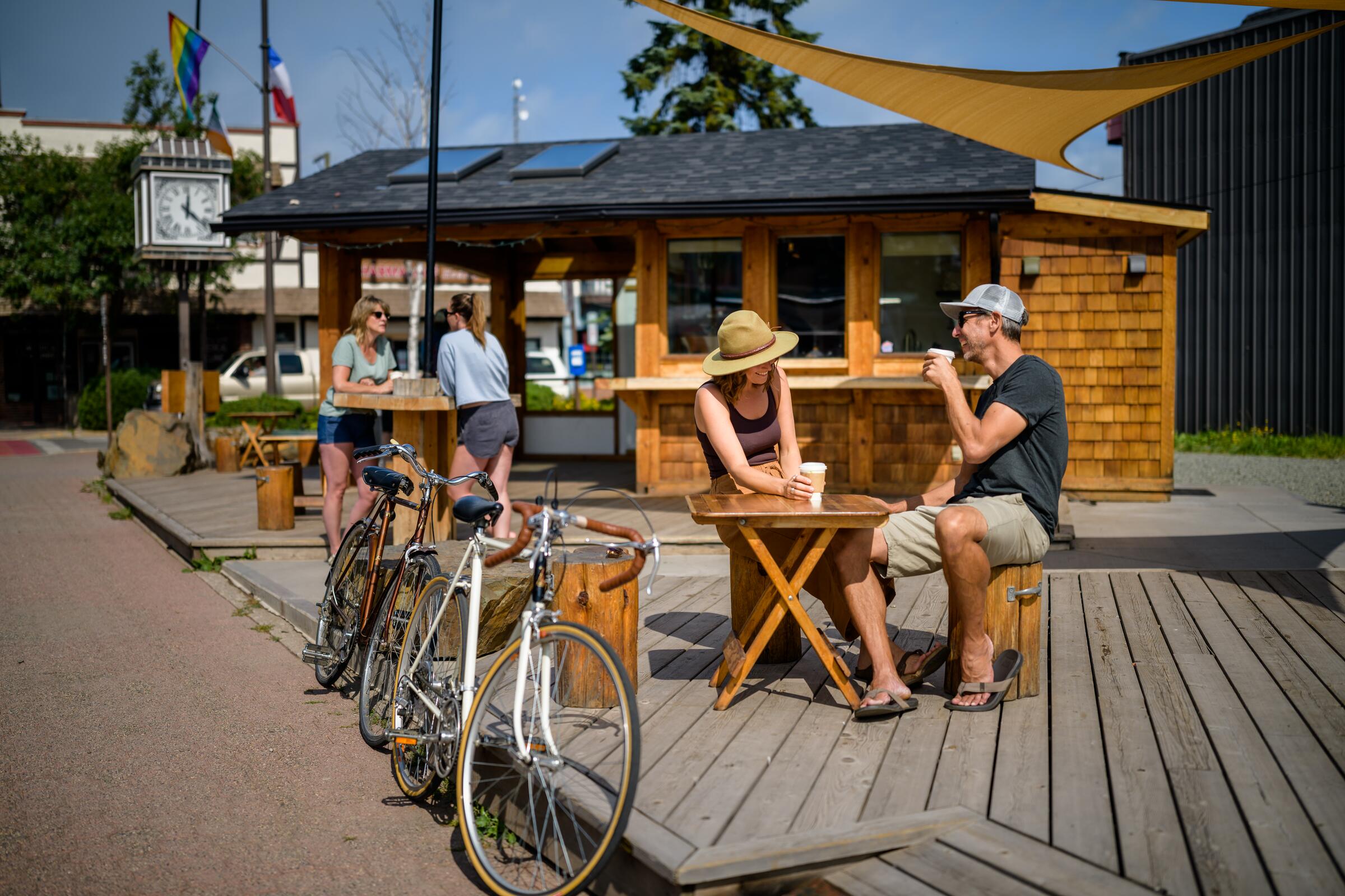 Two people having coffee outside a wooden kiosk cafe, with touring bikes leaned alongside the deck