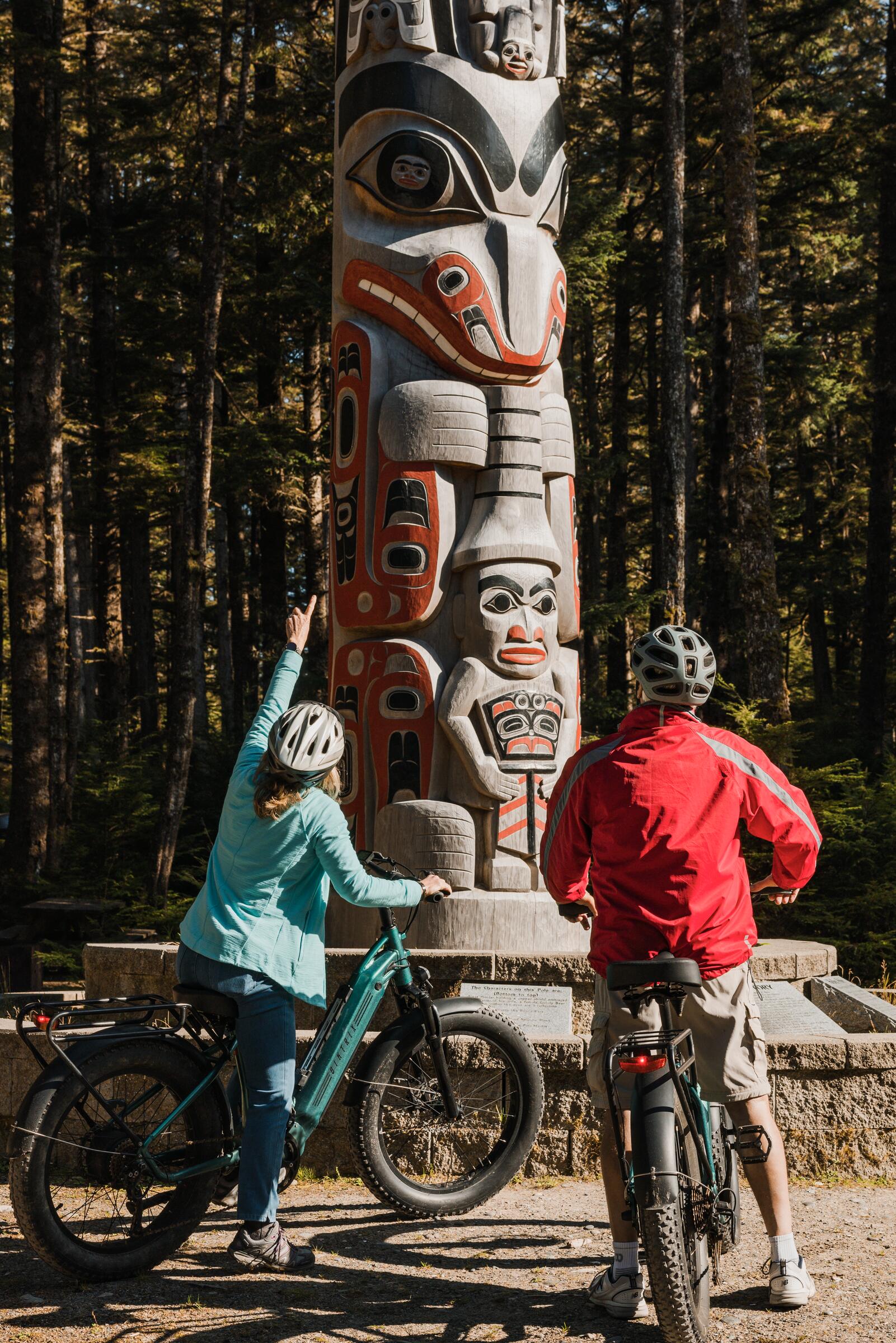 Two riders on e-bikes look up at a carved pole by master carver Christian White at Hiellen Longhouse Village